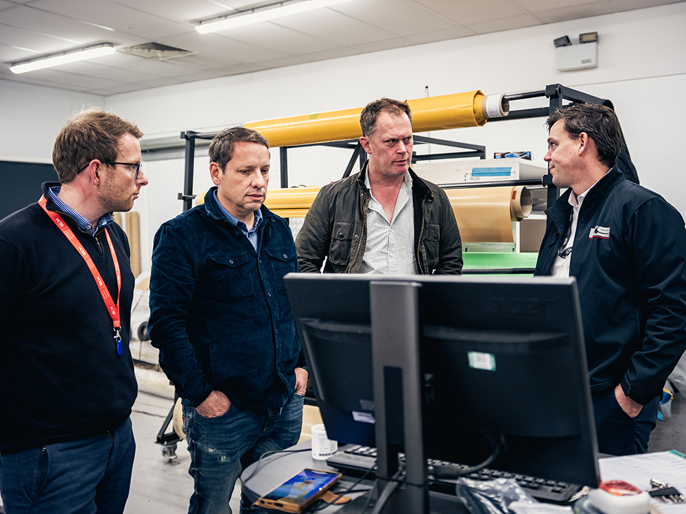 Jake Cronk (COO), Jonathan Wilson (CEO), Charles Fawcett (founder) and Jamie Smith (MD) in a composites workshop reviewing designs beside carbon fibre materials and production equipment.