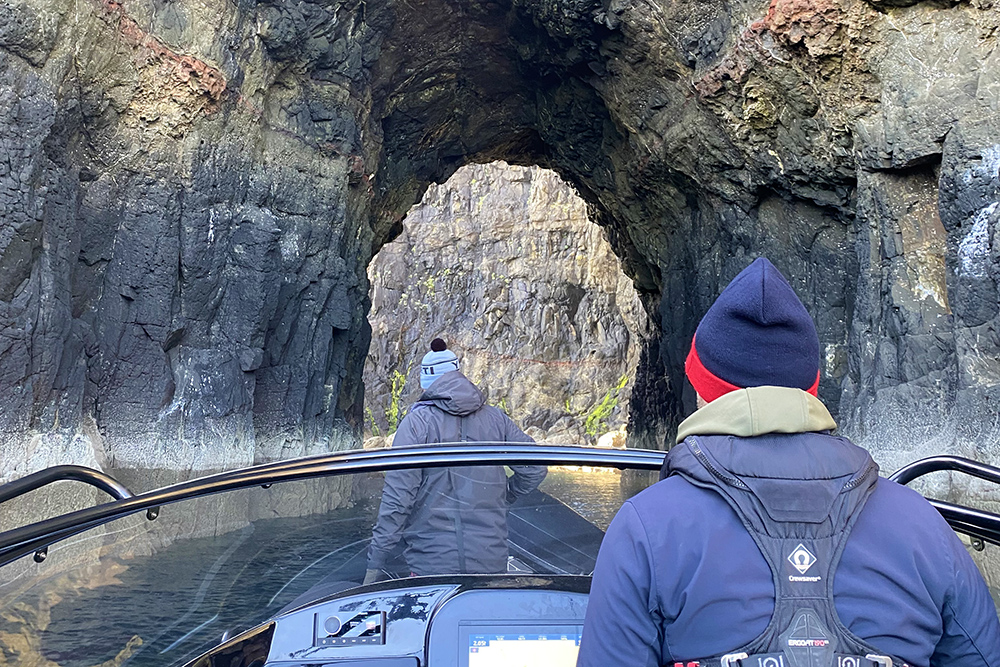 Two team members on a RIB steering toward a volcanic rock cave passage, surrounded by steep dark cliffs and calm water.