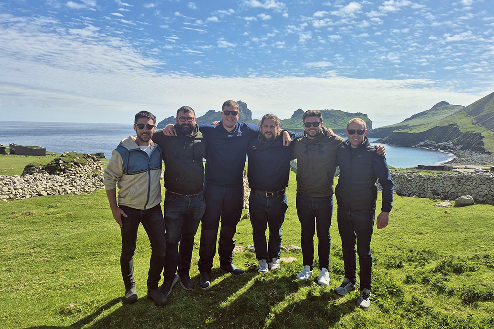 Six friends standing together on a grassy coastal hillside, with blue sea and rugged cliffs in bright summer weather.
