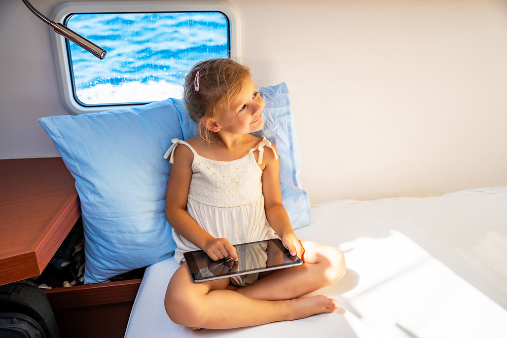 Little girl sits with tablet near window in the cabin of a boat during a sea crossing.