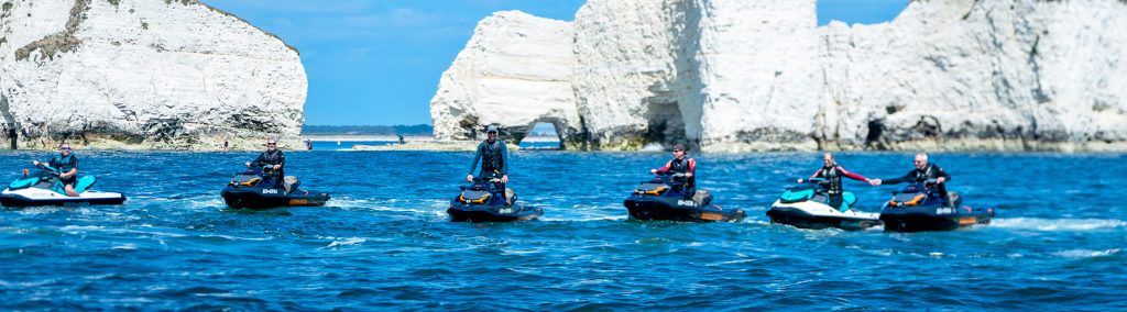 Group of riders on Sea-Doo jet skis cruising in formation past the white chalk stacks of Old Harry Rocks on a bright, calm day.