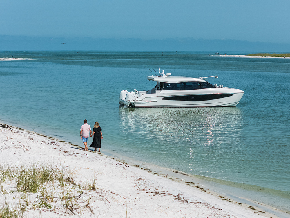 Couple walking along a sandy shoreline beside a moored Aquila 42 Coupe in calm blue coastal water.