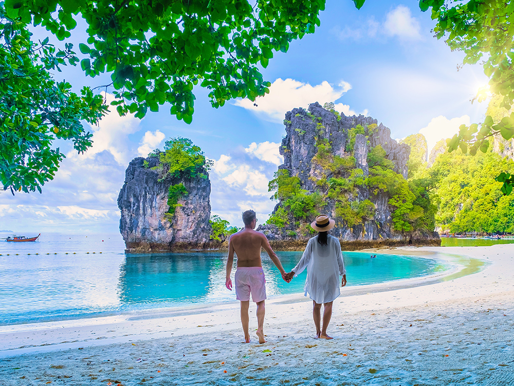 Couple holding hands on a beach, facing turquoise water and limestone cliffs framed by green foliage in bright sunshine.