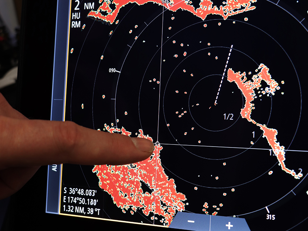 Finger pointing at a Simrad radar display showing coastline returns, range rings and target echoes for navigation awareness.