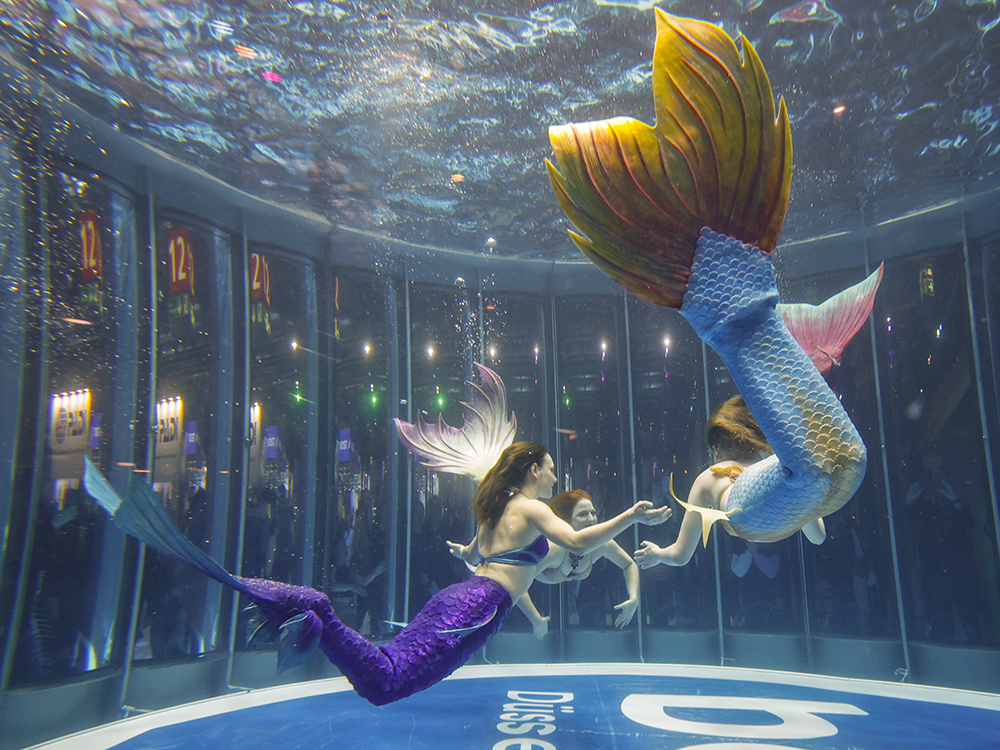 Performers in colourful mermaid tails swim in a large aquarium tank at boot Düsseldorf, entertaining visitors gathered outside the glass.