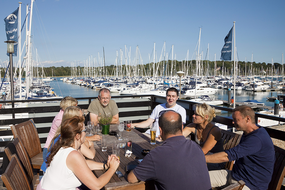 Group enjoying drinks at MDL's Mercury restaurant terrace, overlooking a busy yacht harbour and moored sailboats on a clear day.
