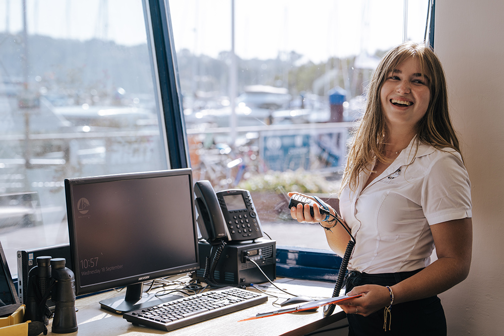 Marina office team member smiling at reception desk with radio in hand, overlooking berths and providing friendly customer support.