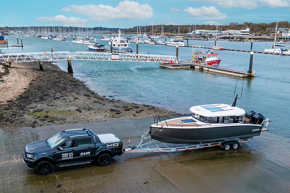 Pickup truck launching a trailer boat on a marina slipway, with pontoons and moored yachts visible across the harbour.