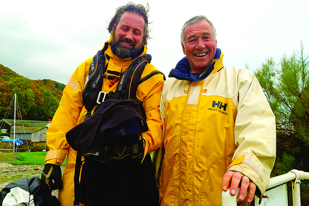 Two smiling sailors in yellow waterproof jackets by a harbour, with autumn trees and boats in the background.