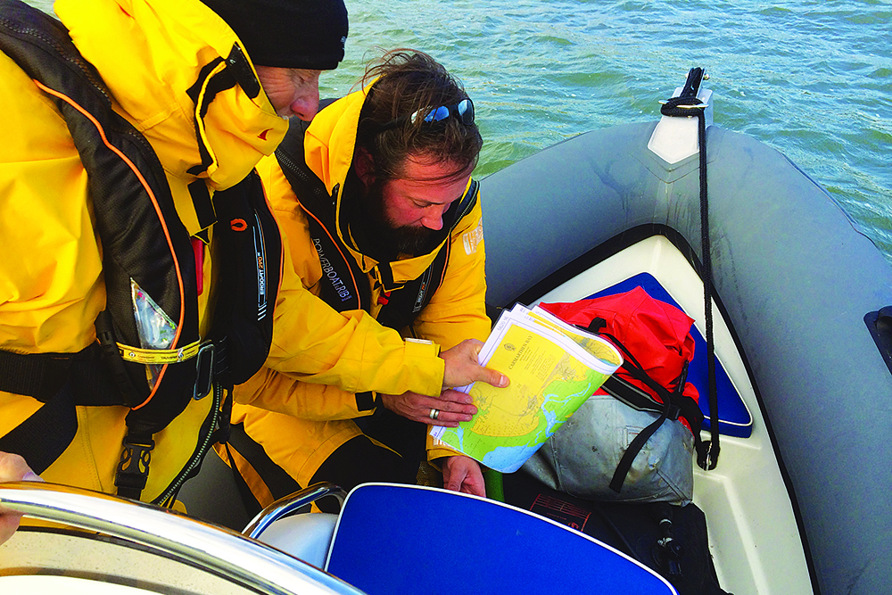 Two crew in yellow foul-weather gear study a paper nautical chart aboard a RIB at sea.