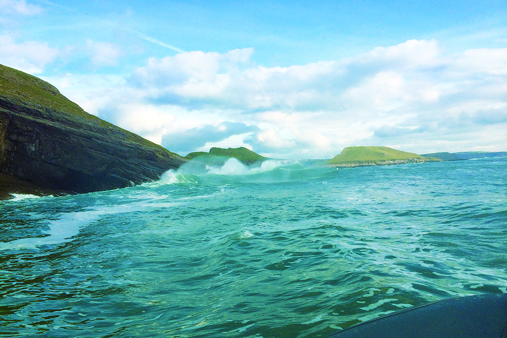Breaking waves surge against rocky cliffs as a RIB runs close along a rugged coastline under blue skies.