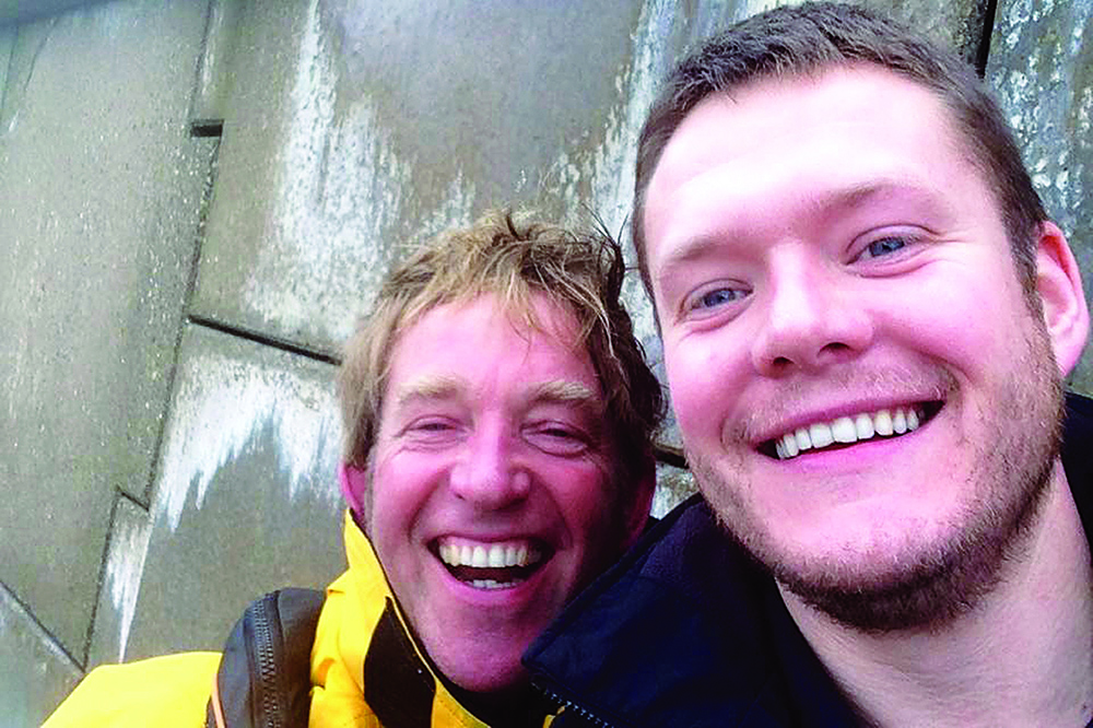 Two smiling men pose for a harbour-side selfie against a stone quay wall.