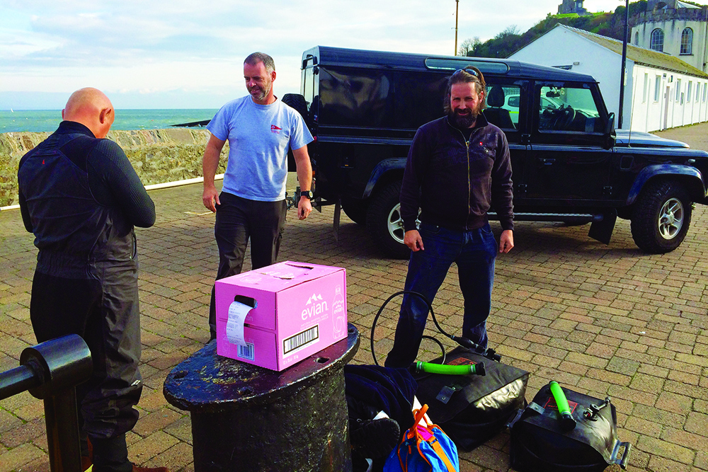 Crew unload gear beside a Land Rover at a harbour quay before departure, sea and harbour wall behind.