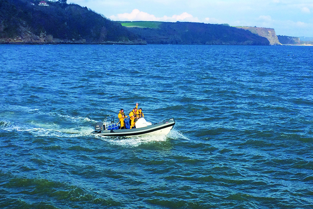 RIB with crew in yellow foul-weather gear runs offshore in Carmarthen Bay near Tenby cliffs.