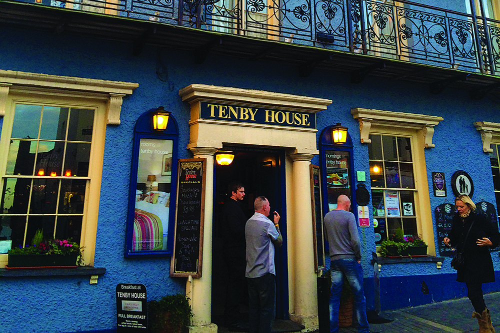 Tenby House pub frontage with patrons outside beneath wrought-iron balcony in Tenby town centre.