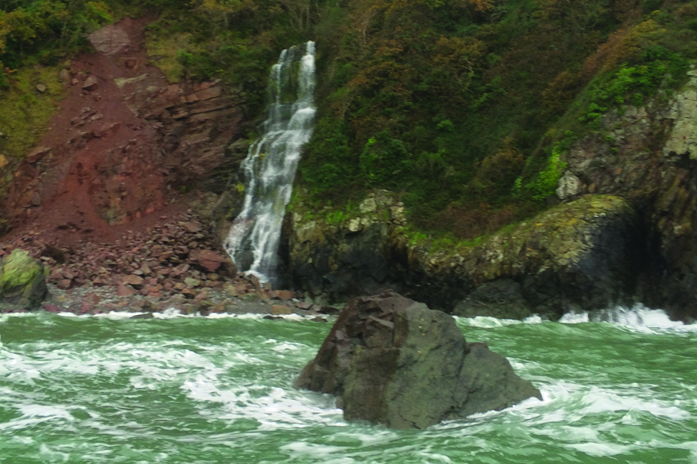 Waterfall cascades down wooded cliff to rocky shore as waves surge below.