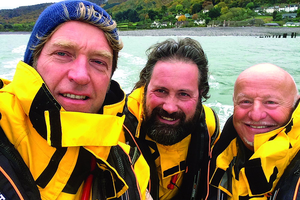 Three smiling crew in yellow foul-weather jackets take a selfie aboard a RIB off the coast.