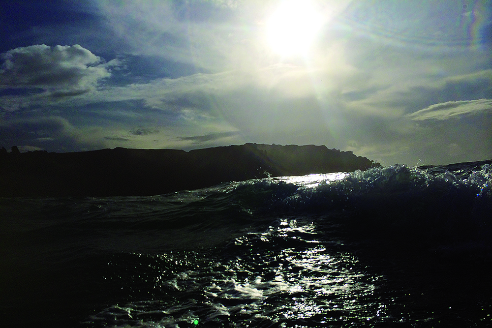 Sunlit waves curl astern near a dark, rocky headland under dramatic skies at sea.