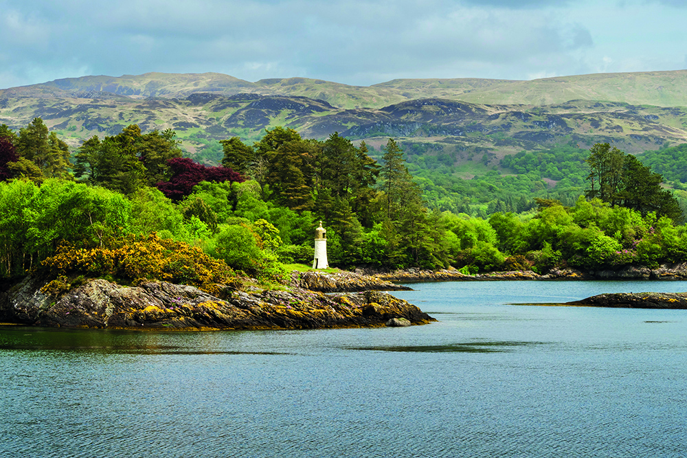 Small lighthouse on rocky shoreline with wooded hills and calm coastal waters in western Scotland.