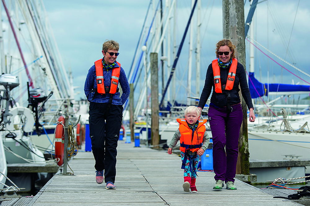 Family in lifejackets walking along marina pontoon with moored yachts at Troon Yacht Haven.