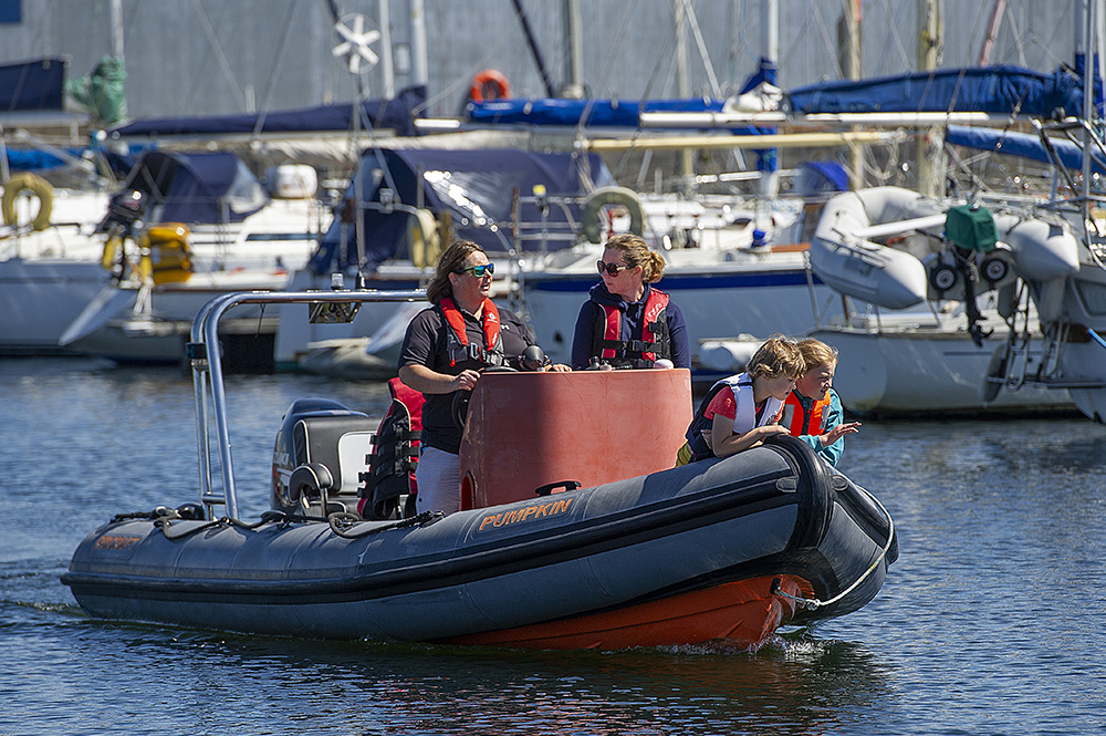 Family in RIB with lifejackets cruising through Troon marina past moored yachts.
