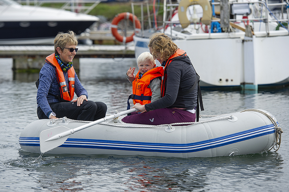 Family in lifejackets rowing small inflatable dinghy in Troon marina among moored yachts.