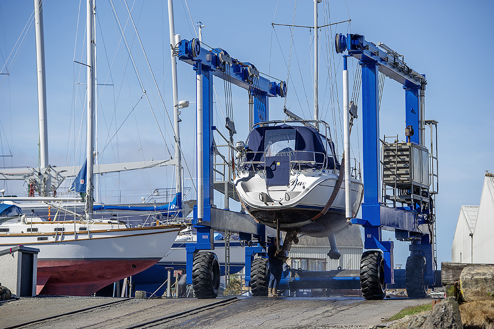 Travel hoist lifting yacht in Troon boatyard with engineer working below and sailboats ashore.