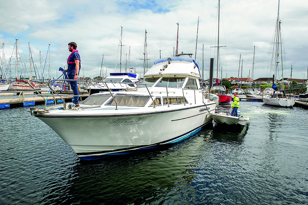 Motor cruiser manoeuvring in Troon marina with crew on deck and tender alongside among moored yachts.