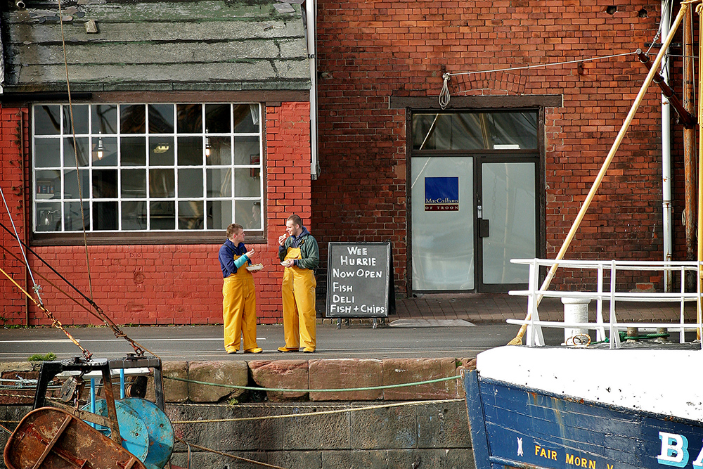 Fishermen in yellow oilskins eating by Wee Hurrie on Troon harbour quay beside moored boat.