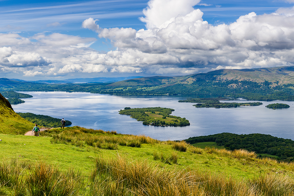 Panoramic view of Loch Lomond with islands, rolling hills and hikers overlooking Scotland’s iconic freshwater lake