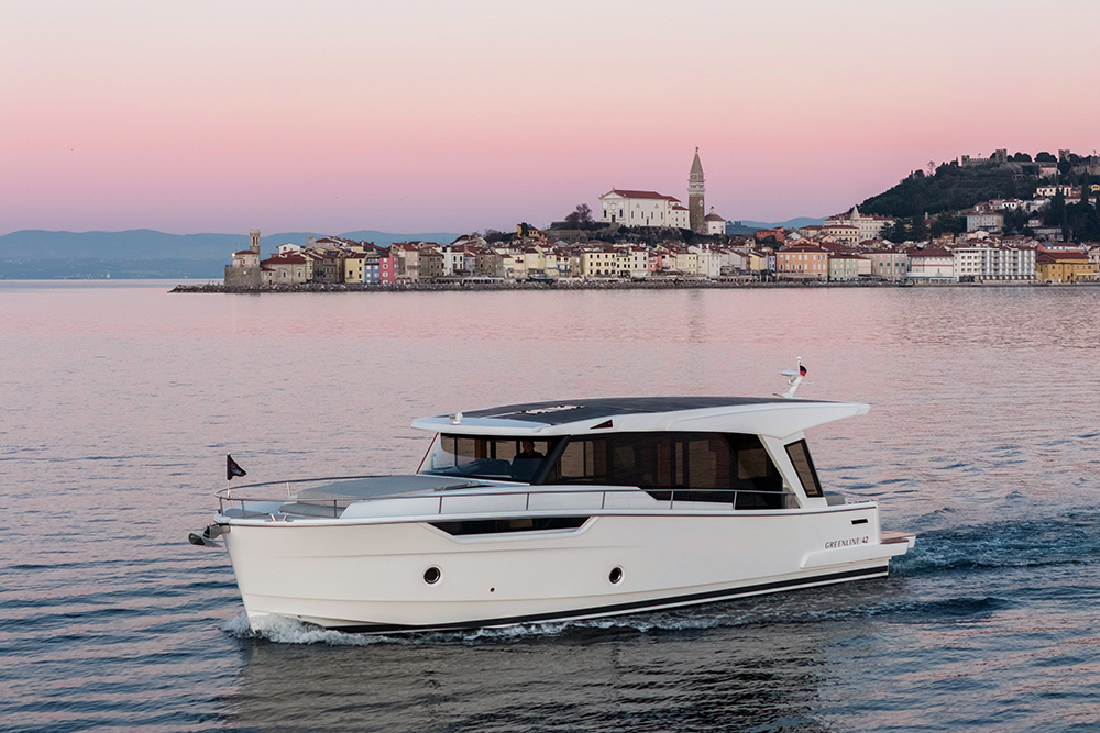 Greenline 42 hybrid motor yacht underway at sunset with coastal town skyline in background.