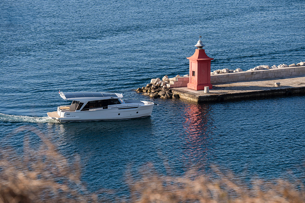 Greenline 42 hybrid yacht cruising past a red harbour lighthouse along a stone breakwater.