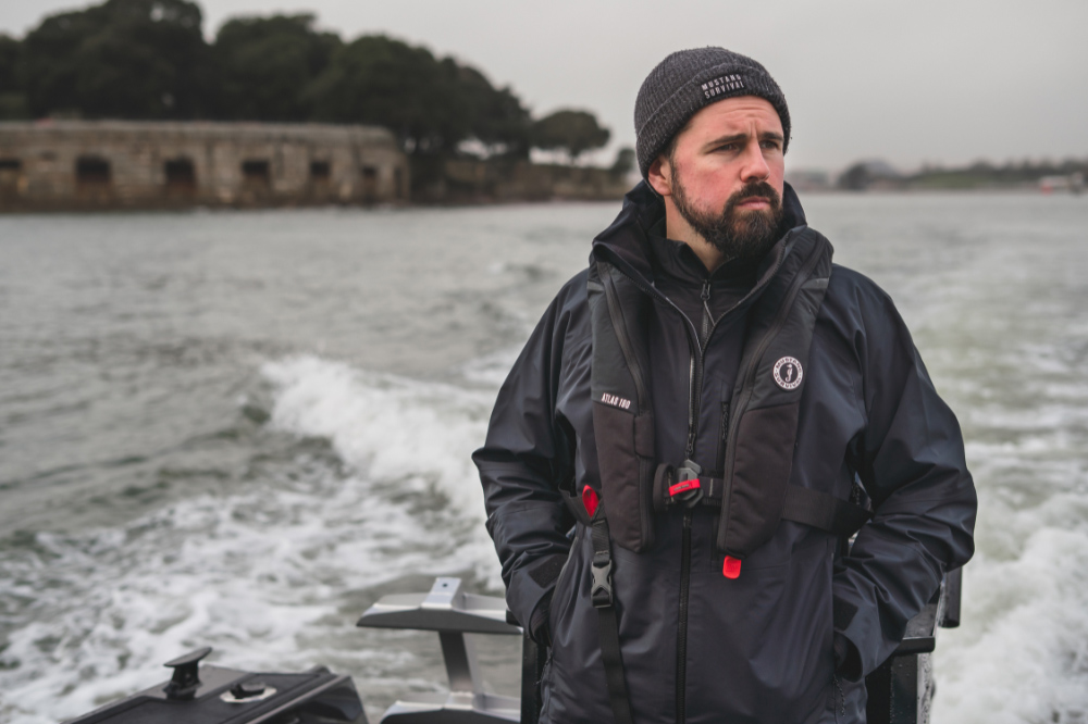 Man wearing Mustang Survival waterproof jacket and life vest aboard a RIB in wet coastal conditions.