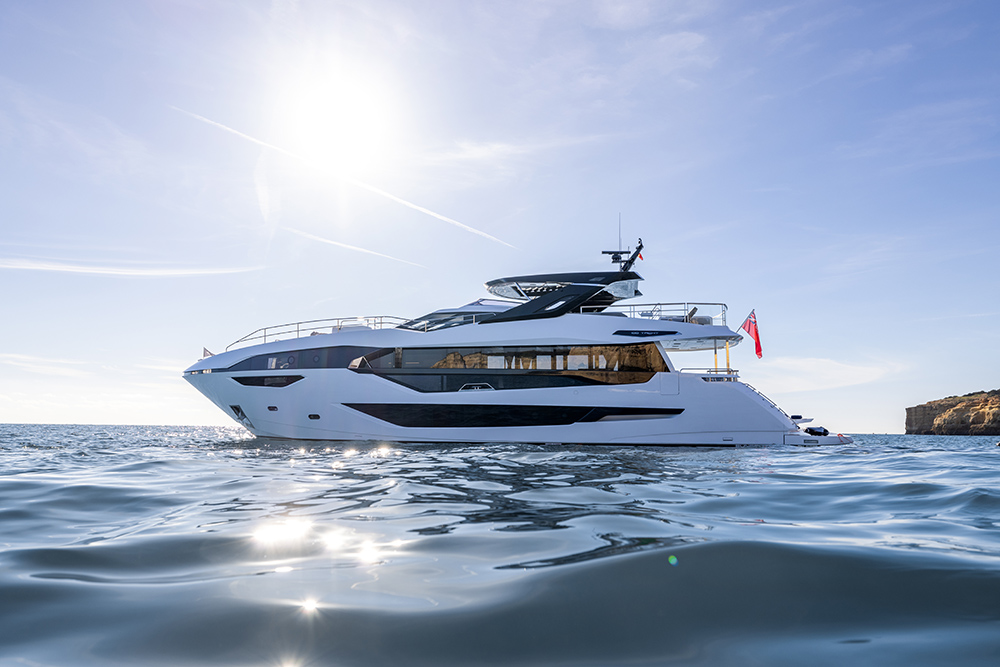 Sunseeker 90 Ocean luxury motor yacht cruising on calm sea near coastline, showcasing sleek profile and expansive glazing.