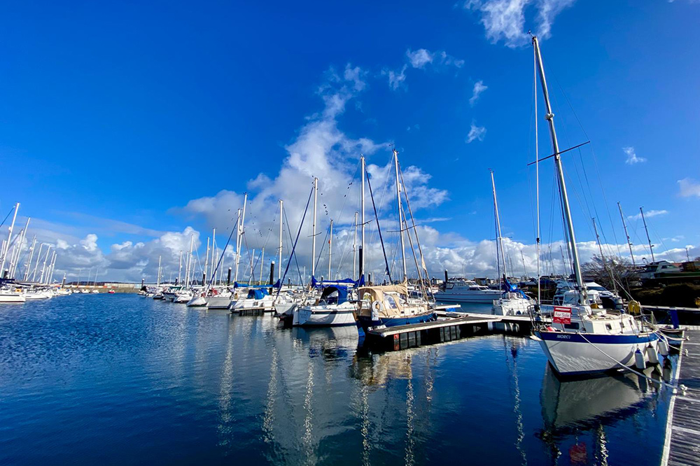 Sailing yachts moored on pontoons at Troon Yacht Haven with calm water and blue sky reflections.