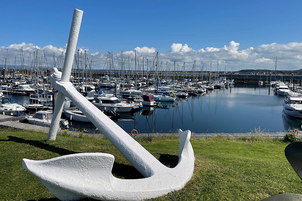 Large white anchor sculpture overlooking Troon marina with moored boats and harbour under clear blue skies.