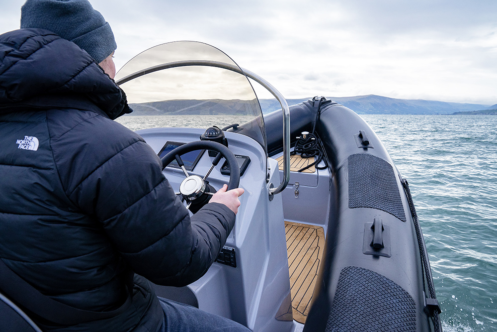 Driver at helm of Redbay Stormforce 650 RIB cruising offshore, with console, tubes and coastal hills in background