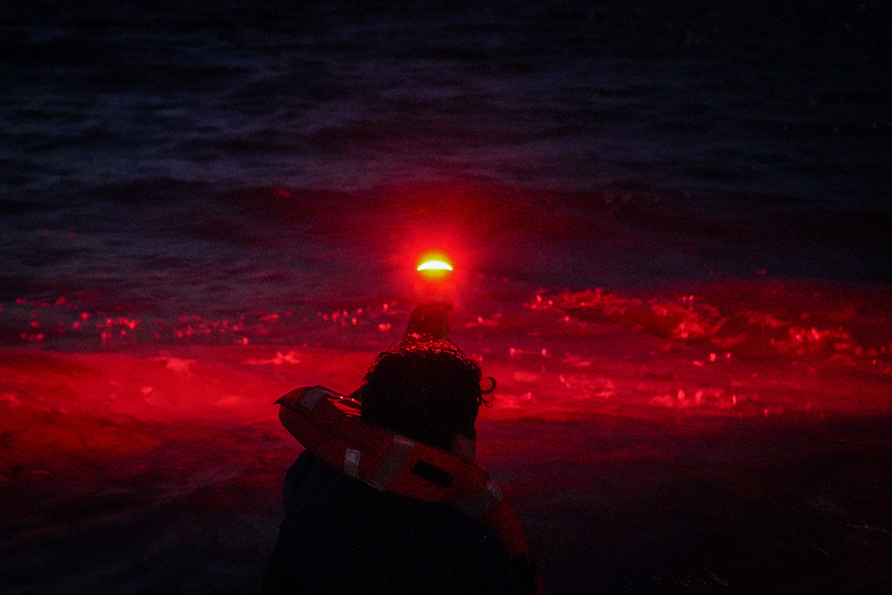A dark night with a man holding up a electronic flare giving a red glow into the sea