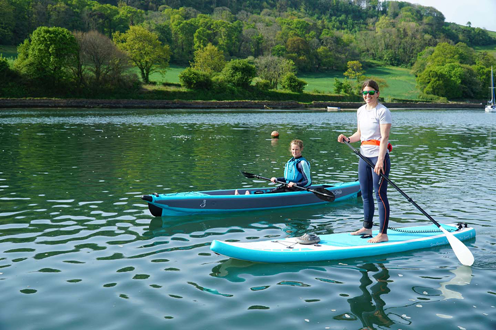 Paddleboarder and child kayaking on calm river with wooded shoreline, enjoying family watersports in sheltered conditions