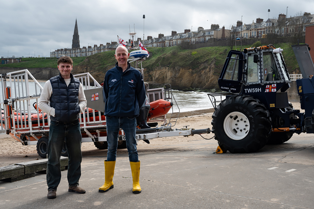 Two men beside an RNLI lifeboat and launch tractor at the coast.