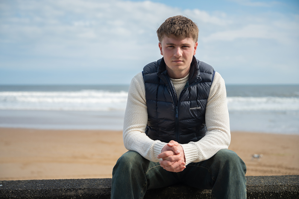 Young man sitting on a beach wall with surf behind.