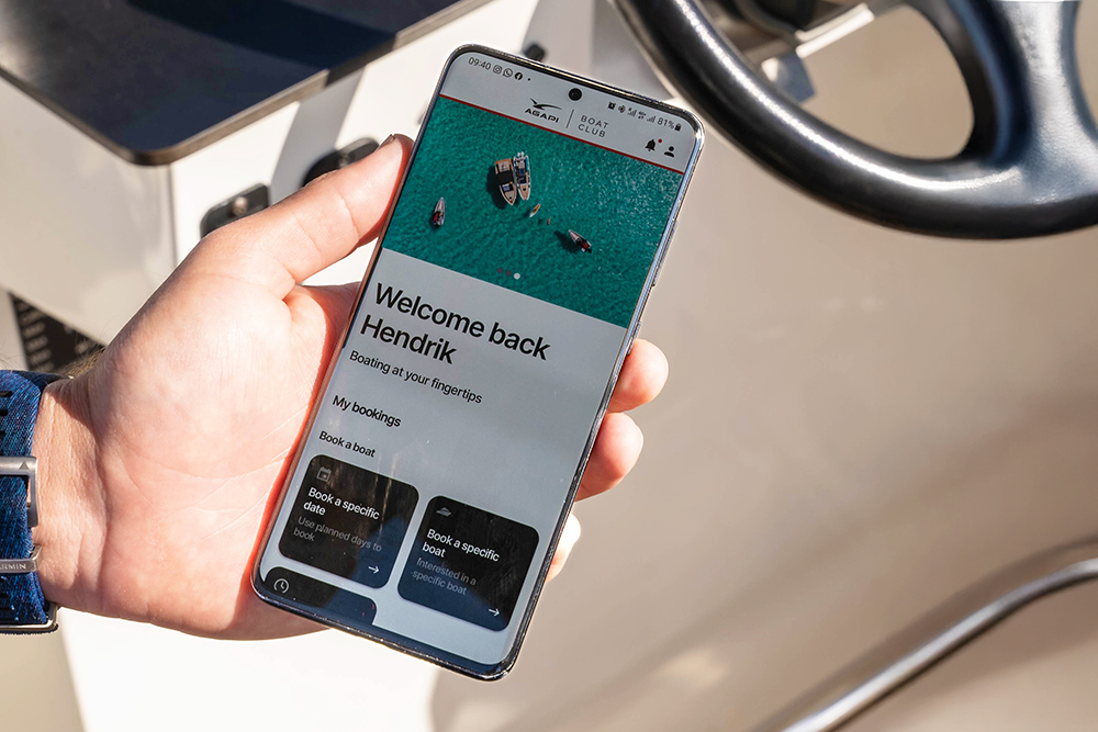 Hand holding Agapi Boat Club app on a boat helm, showing digital booking for shared Axopar boating