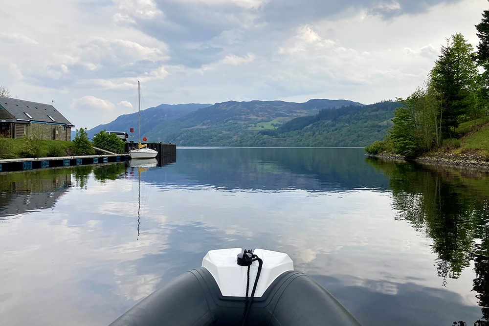 RIB bow approaching Loch Ness on calm water, with mountains and forest reflected ahead