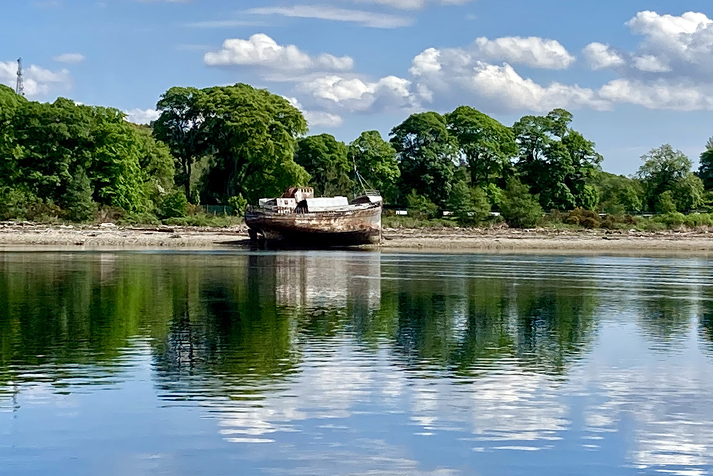 Corpach wreck on Loch Linnhe reflected in calm water, with woodland along the Highland shore