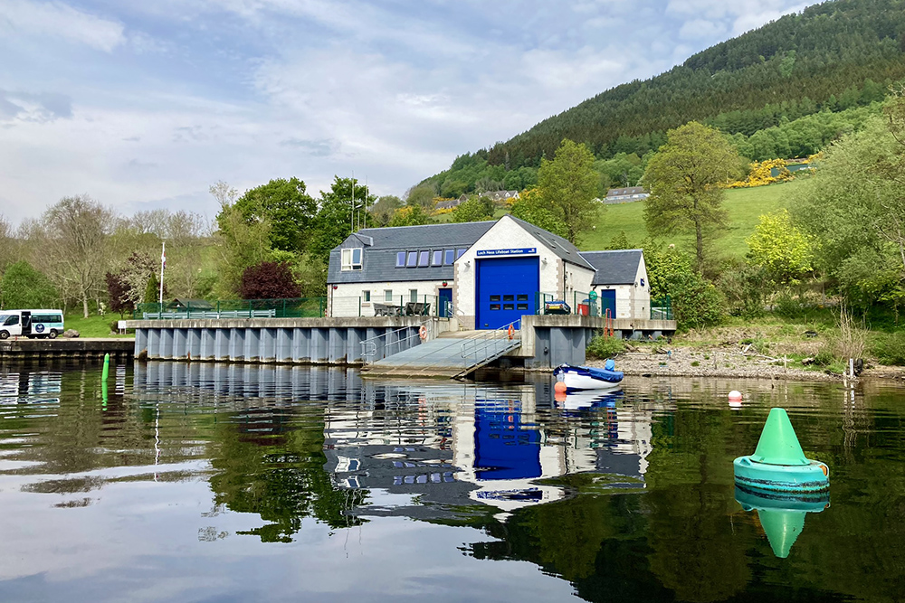 Loch Ness RNLI lifeboat station reflected in calm water beneath wooded Highland slopes