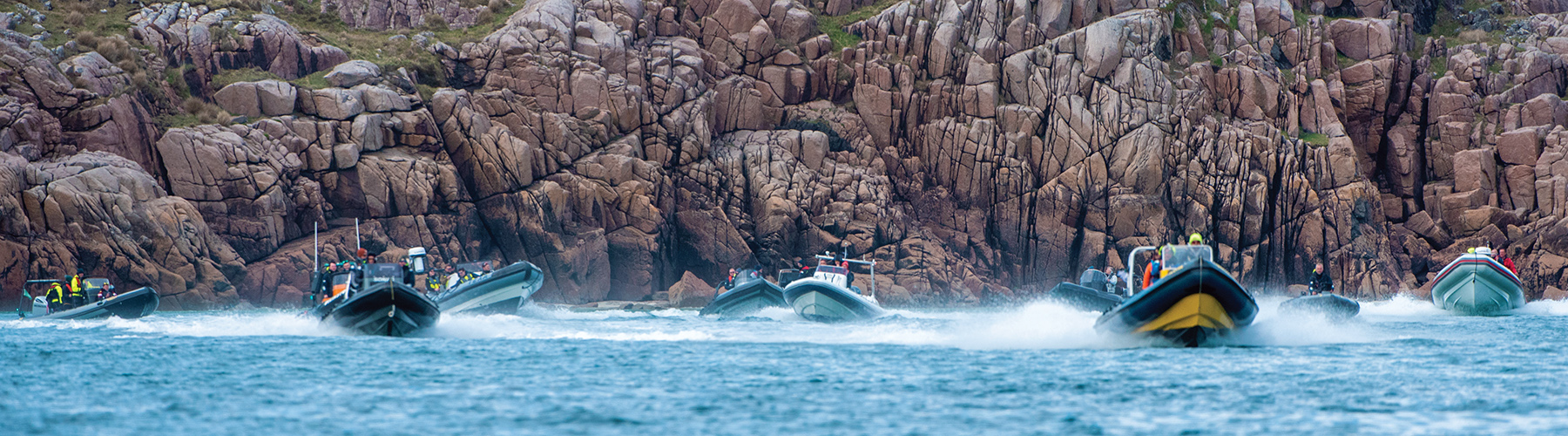 Multiple rigid inflatable boats underway underneath a cliff face