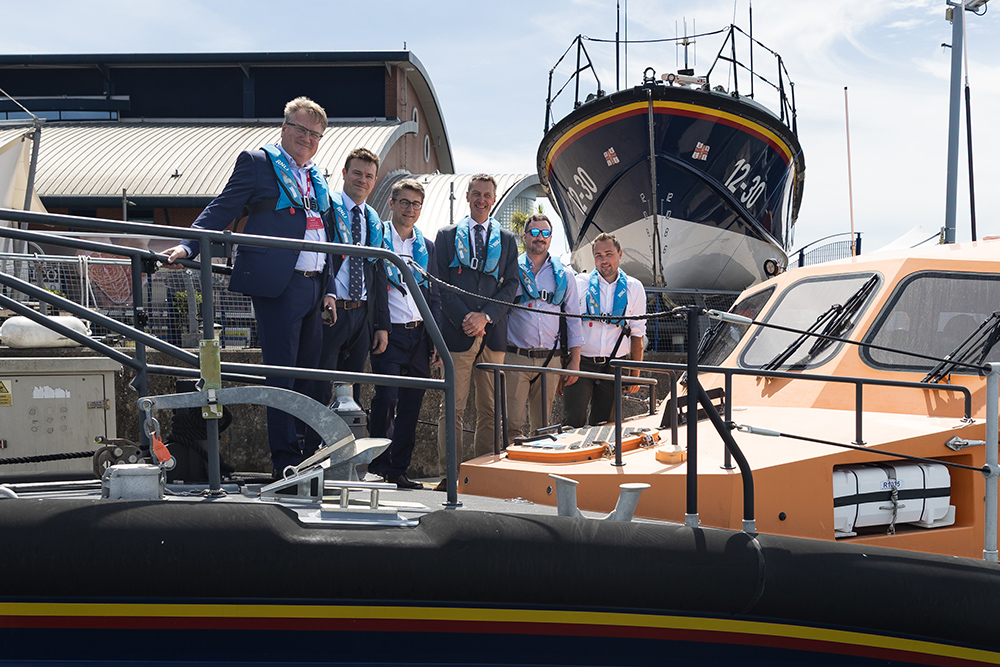 RNLI and Raymarine representatives in lifejackets beside an inshore lifeboat, with a larger lifeboat behind.