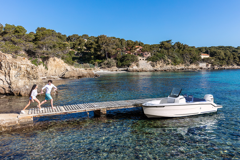 Jeanneau Cap Camarat 6.0 CC moored beside a wooden jetty as a couple runs towards it in a rocky cove.