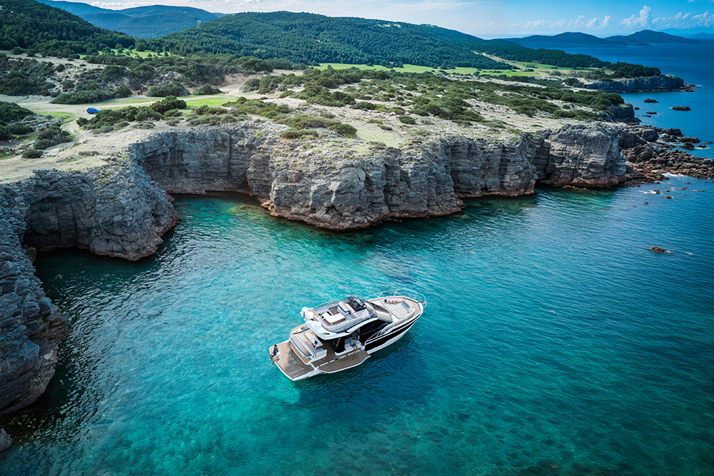 Aerial view of a Galeon flybridge yacht anchored in a turquoise cove, framed by limestone cliffs and forested coastline.