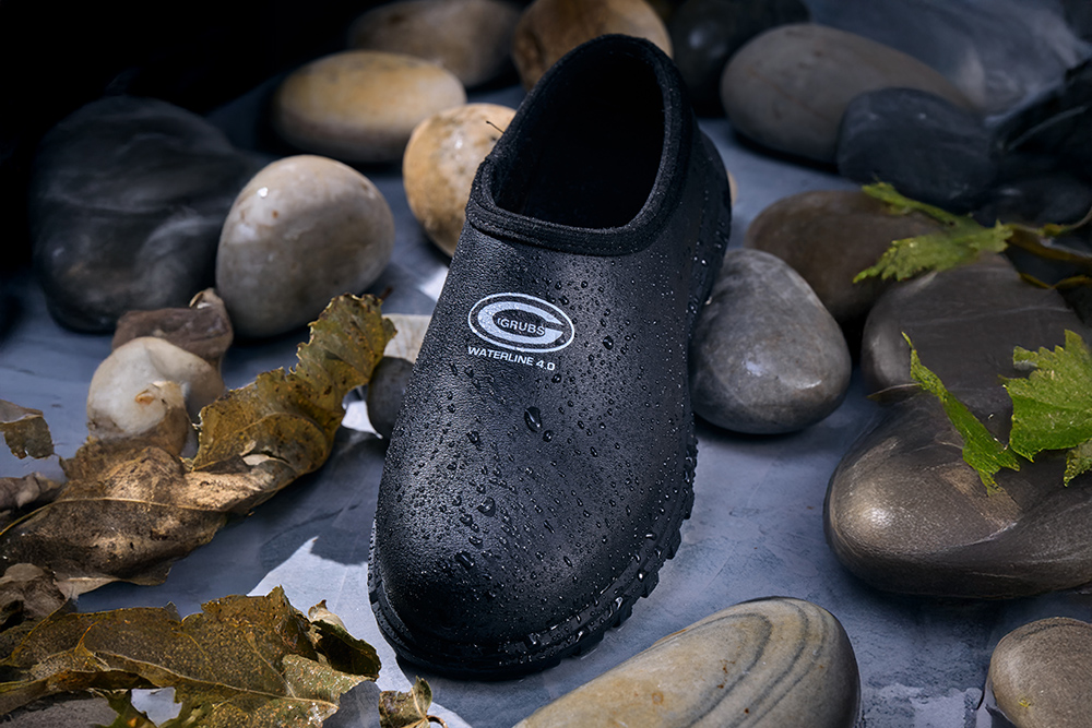 Grubs Waterline waterproof slip-on shoe with water droplets, displayed among wet rocks and leaves.